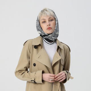 Silk scarf with elegant black and white pattern worn as a headscarf with a beige trench coat and white sweater, showcasing classic styling