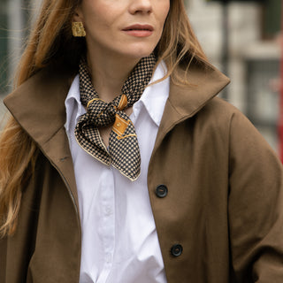 Woman wearing a black and beige houndstooth silk scarf with brown coat, white shirt in a blurred background.