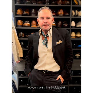 Stylish blue and red silk neckerchief worn with dark suit and cream shirt, displayed against a backdrop of elegant footwear