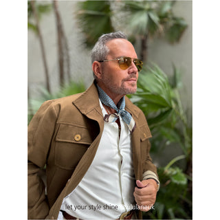 Person wearing blue patterned silk bandana as neckerchief with brown jacket and white shirt against tropical foliage background