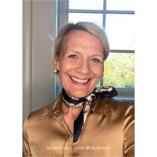 A luminous purple blue with white print silk scarf is the focal point, gracefully tied around the neck of a blonde woman in her fifties. The scarf’s rich tones and fluid drape bring a refined pop of colour and elegance to her classic look.