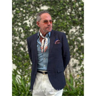 Vintage baroque silk scarf styled as a neckerchief with navy blazer, denim shirt, and white trousers against green foliage backdrop