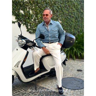 Stylish person on white scooter wearing denim shirt and vintage baroque silk scarf with white pants against ivy wall backdrop