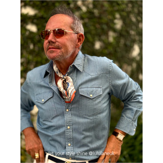 Person wearing a light blue denim shirt styled with a vintage baroque silk scarf in cream, blue and orange tones tied loosely at the neck
