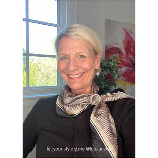 A refined black and beige houndstooth silk scarf takes center stage, elegantly wrapped around the neck of a blonde woman in her fifties and fastened with a delicate pearl pin, adding a sophisticated touch to her look.