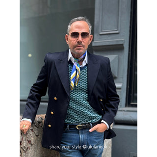 Stylish person wearing blue and yellow silk scarf as neck accessory with navy blazer, green patterned sweater and jeans in urban setting