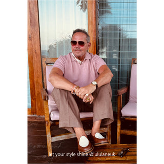 Person sitting in wooden chair wearing pink polo shirt and neutral pants, styled with sunglasses and accessories in tropical setting