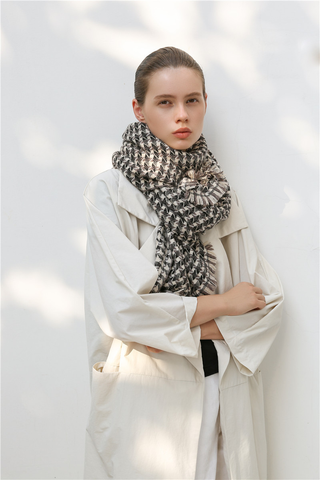 Woman wearing a taupe, black and cream white cashmere scarf with houndstooth check pattern and white coat against a light background
