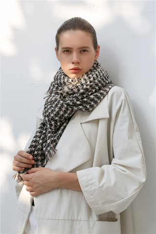 Woman wearing a taupe, black and cream white cashmere scarf with houndstooth check pattern and white coat against a light background