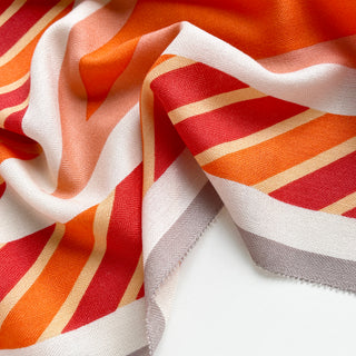 Close-up of a diamond-shaped silk cashmere wool scarf in orange, red, taupe, and cream with bold geometric stripes.