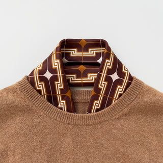 Retro 70s pattern silk scarf in burgundy, amber and beige with geometric pattern, styled with a tan sweater, showcasing hand-rolled edges