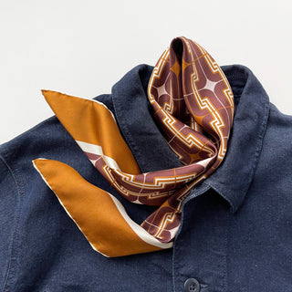 Retro 70s silk scarf in burgundy, amber and beige geometric pattern draped on dark blue denim shirt collar, showcasing hand-rolled edges