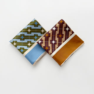 Two folded men's luxury silk square scarves featuring retro geometric pattern in warm burgundy brown and dusty blue & olive green colour ways. 