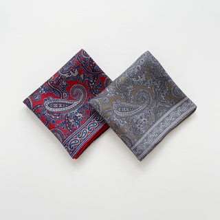 Two folded paisley patterned men’s silk neckerchief scarves, one red and navy, the other taupe and baby blue, on a light grey background.