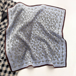 Pale blue silk bandana scarf with intricate paisley pattern, brown border, and delicate detailing, displayed with elegant draping on checkered fabric