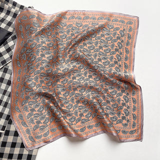 Square silk bandana scarf in coral pink with navy blue paisley pattern throughout and decorative border, displayed with gentle folds against checkered fabric