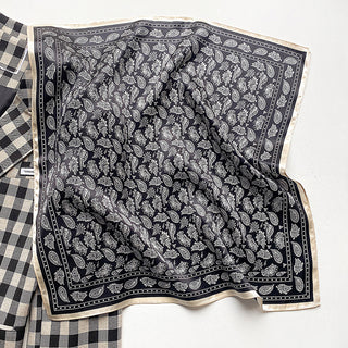 Black silk bandana with white paisley pattern and cream border, displayed on checkered fabric showing luxurious texture and intricate design