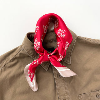 Red silk scarf with white crown pattern tied as a neckerchief around the collar of a khaki button-up shirt, featuring a pink border edge