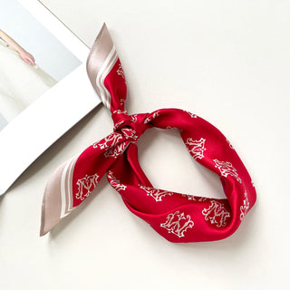 Red silk square scarf with white crown pattern and beige border, styled in a knotted bandana form on light background