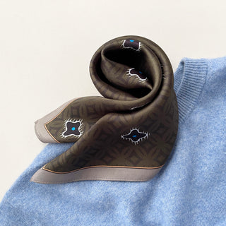 Folded olive green silk neckerchief with geometric star pattern featuring blue and white accents, elegantly draped on light blue knitwear