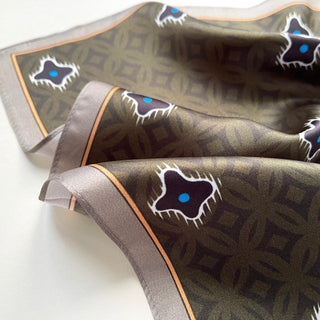 Olive green silk scarf with geometric pattern featuring black stars with blue centers, bordered with gray and gold trim, displayed with elegant folds