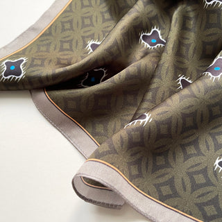 Olive green silk neckerchief with geometric pattern, featuring black star motifs with blue centers, bordered by a soft gray trim with gold accent line.
