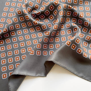 Close-up of a lead grey silk square scarf by LuluLane with rust red four-petal flower motif, double-sided print and hand-rolled hems.
