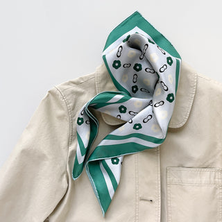 Square silk scarf with teal green border and simplified floral pattern on light grey background, artfully draped on beige jacket collar