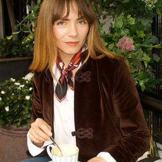 Woman wearing a burgundy red patterned silk bandana scarf with brown velvet coat and white shirt, sitting among greenery.