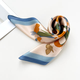 Square silk scarf with blue border featuring orange iris flowers, green leaves, and butterfly motif on champagne background, elegantly draped
