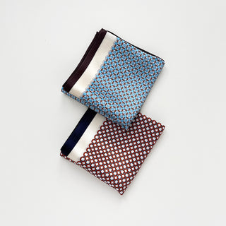 Two folded silk scarves with geometric lattice patterns - sky blue with brown edge (top) and red with navy edge (bottom) on white background