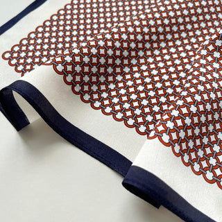 Luxury silk scarf with red geometric lattice pattern on cream background, featuring navy blue border trim, showcasing smooth fabric texture