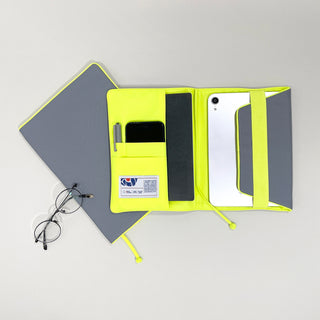 Eco-friendly notebook cover in grey with neon yellow interior, featuring pockets for tablet, phone, and pen, displayed open with a notebook and glasses