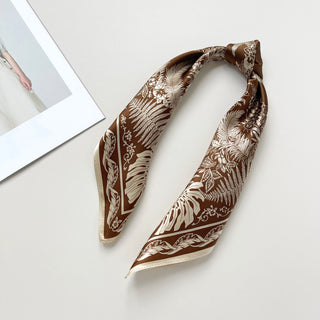 Bronze silk bandana scarf with elegant white botanical pattern featuring fern and monstera leaves, displayed on white surface with cream border.