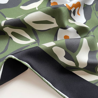 Close-up of a silk bandana scarf by LuluLane with green background, cream white floral print, and yellow leaves, framed by black border with hand-rolled edges.