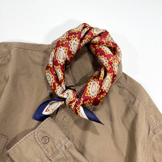 Red silk neckerchief with paisley pattern in gold and white, styled around collar of tan shirt, featuring navy blue accent on edge