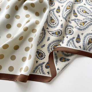 Close-up of Beige silk scarf by LuluLane with intricate black paisley pattern on one side, solid cream colour on the other, featuring elegant brown border trim