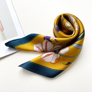Mustard yellow silk scarf with floral and butterfly pattern, featuring white flowers, purple accents, and teal border, elegantly arranged showing luxurious sheen.
