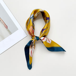 Mustard yellow silk bandana scarf with floral and butterfly print, featuring navy blue trim and tied in classic knot style on white surface