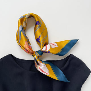 Mustard yellow silk bandana scarf with floral and butterfly print, knotted stylishly around neckline of navy top, showcasing teal border