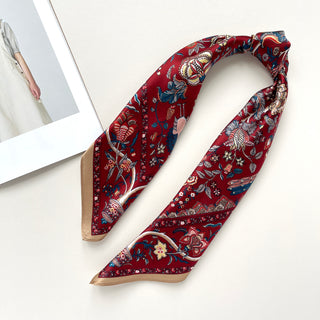 Rich red silk scarf with vintage floral garden pattern featuring blue, gold and pink botanical designs with beige trim, displayed on white surface