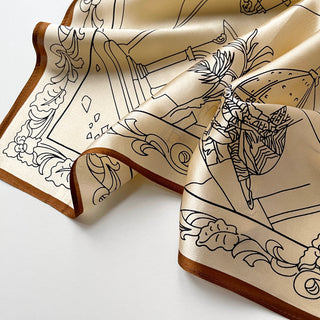 beige silk scarf with black line art illustrations and copper-brown border trim, elegantly draped to display its luxurious sheen and intricate design