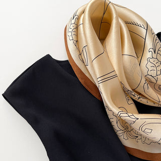 Beige silk bandana scarf with elegant black line art design and cognac border, displayed alongside a black fabric on white background