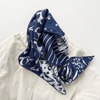 Navy and white silk scarf with mixed patterns including floral, polka dots, and animal prints, elegantly draped on a light-colored shirt.