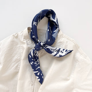 Navy Urban Safari silk scarf with white patterns tied around collar of cream shirt, featuring mixed floral, polka dot, and animal print motifs