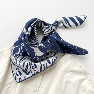 Navy and white silk scarf featuring mixed patterns of floral, polka dot, leopard print and stripes, styled as a neckerchief on light collar
