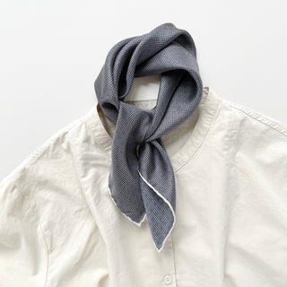 Navy blue silk scarf with white grid pattern tied elegantly around collar of cream shirt, showing white edge trim and luxurious drape