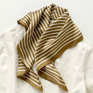 Luxurious bronze and white striped silk bandana scarf draped elegantly over a cream blouse, showcasing its shiny texture and geometric pattern.