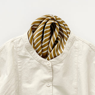 Golden brown silk scarf with diagonal white stripes elegantly styled around a cream blouse collar, showcasing luxurious sheen and texture