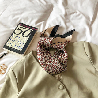 Folded red and black geometric silk bandana scarf with elegant pattern displayed on white bedding next to beige blazer and book